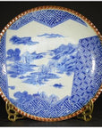 Blue and white ceramic Igezara transferware plate with landscape and geometric rim, on metal stand.