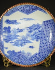 Blue and white ceramic Igezara transferware plate with landscape and geometric rim, on metal stand.