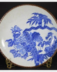Blue and white ceramic Igezara transferware plate with floral and landscape designs on brass stand, black background.