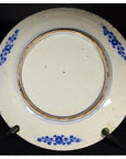 Blue and white porcelain plate with floral rim on black stand, dark background.