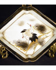 White porcelain Kutani dish with hand painted cranes, pine trees, and gold accents on a display stand.