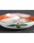 Close-up of antique Japanese Satsuma ceramic plate with green leaf, red dot, and orange rim on dark background.