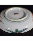 Close-up of an antique Japanese Satsuma porcelain plate with green leaves and red designs on a dark surface.