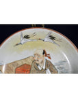 Close-up of an antique Satsuma porcelain plate with painted elderly man and two flying cranes on dark background.