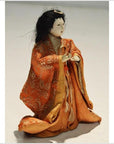 Orange kimono-clad antique Japanese Taisho Samurai doll, kneeling with raised hands and serene expression, close-up.