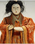 Close-up of antique Japanese Taisho samurai doll with white face, black hair, and ornate orange-gold kimono.