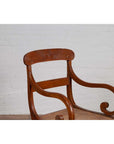 Brown wooden and rattan Javanese armchair with carved back and curving arms against a white brick wall indoors.