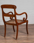 Brown wood and rattan armchair with carved back and curving arms on wood floor next to white brick wall indoors.