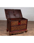 Brown hand carved wooden blanket chest with gilt accents, open lid, on wood floor against white brick wall.