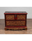 Brown wooden blanket chest with hand-carved details and gilt accents on hardwood floor against a white wall.