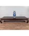 Dark wood opium bed with carved legs against a white brick wall, antique Ming Dynasty style, indoor setting.