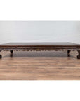 Dark wood ornate carved opium bed in Ming Dynasty style, placed on wooden floor against a white brick wall indoors.