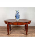 Brown wood oval dining table with spindle legs and warm patina, placed against a white brick wall.