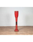 Red wood ceremonial drum with round base on wooden floor in front of white brick wall indoors.