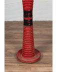 Red wood ceremonial drum with round base on wooden floor next to a white brick wall indoors.