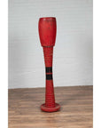 Red wooden ceremonial drum with round base on wooden floor beside a white brick wall indoors.