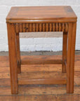 Brown wooden Art Deco stool on wooden floor in front of white brick wall.
