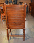 Brown wooden Arts & Crafts style chair with slatted backrest, rear view on wooden floor among other furniture.
