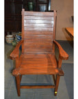 Brown teak armchair with slatted back and seat, Arts & Crafts style, indoors on a patterned floor.