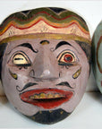 Colorful Balinese mask with uneven eyes, mustache, headband, and wide jagged smile, close-up.