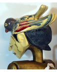 Brown hand-carved wooden puppet with painted bird headdress and stylized features, shown indoors.
