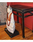Close-up of a Buddha statue with a red bead necklace in front of a black altar table on a wooden floor.