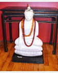 White Buddha statue with orange and red bead necklaces on floor, Black altar table with gold accents in background.