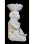 White porcelain monk figurine seated, holding scalloped dish, with visible chips and discoloration, close-up view.
