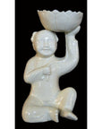 White porcelain monk figurine kneeling and holding bowl aloft, set against a black background.
