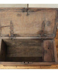 Brown wooden blanket chest with metal hinges, open and empty, sitting on a scratched wooden floor indoors.