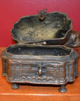 Brass animal-shaped box with legs and hinged lid, open on wood surface, red wall background, close-up.