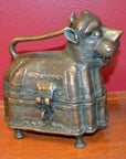 Brass animal-shaped box with handle, latch, and four legs on wooden surface against red wall.