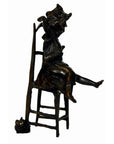 Bronze girl doll sculpture sitting on tall chair with outstretched legs, small cat at base, close-up view.