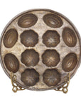 Round bronze shell pan mold with 12 flower and shell shapes, displayed on a metal stand.