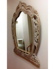 Carved mirror with decorative frame on wall, reflecting hallway, white vase, and part of a chair in view.