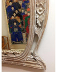Brown carved wood mirror reflecting colorful painting of people in traditional attire on plain white wall.