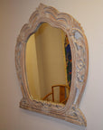 Ornate carved mirror with decorative frame reflecting a chair and colorful wall artwork in the background.