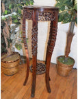 Brown carved wood plant stand with ornate details, placed on a wooden floor between two green potted plants.