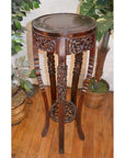 Brown carved wooden plant stand with decorative patterns on a wooden floor next to green potted plants.