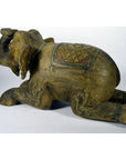 Brown wood hand-carved elephant figurine with decorated saddle, kneeling, trunk raised, close-up on neutral background.
