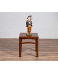 Ceramic figurine in traditional dress on brown Ming style side table, white brick wall background.