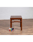 Brown wood Ming style side table with humpback stretcher, antique finish, on wood floor against a white wall.