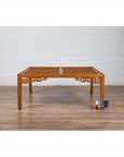 Brown elmwood square coffee table with open fretwork and horse-hoof legs on wood floor by white brick wall indoors.