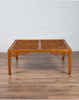 Brown elmwood square coffee table with open fretwork and horse-hoof legs on wooden floor by white brick wall indoors.