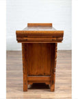 Brown elmwood two-door low cabinet with everted flanges and scrolls, side view, set against a white brick wall.