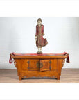 Brown wood two-door low cabinet beside wood table with red cloth and standing Buddha, on wooden floor indoors.