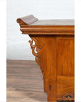 Brown elmwood low cabinet with everted flanges and scrolls, indoors against a white brick wall and wooden floor.