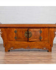 Brown elmwood two-door low cabinet with everted flanges and scrolls, indoors against a white brick wall.