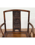 Brown carved elmwood chair with animal and floral backrest designs, positioned indoors by a white brick wall.