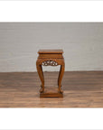 Brown wooden Ming style incense stand with cabriole legs and carved apron, placed next to a white brick wall indoors.